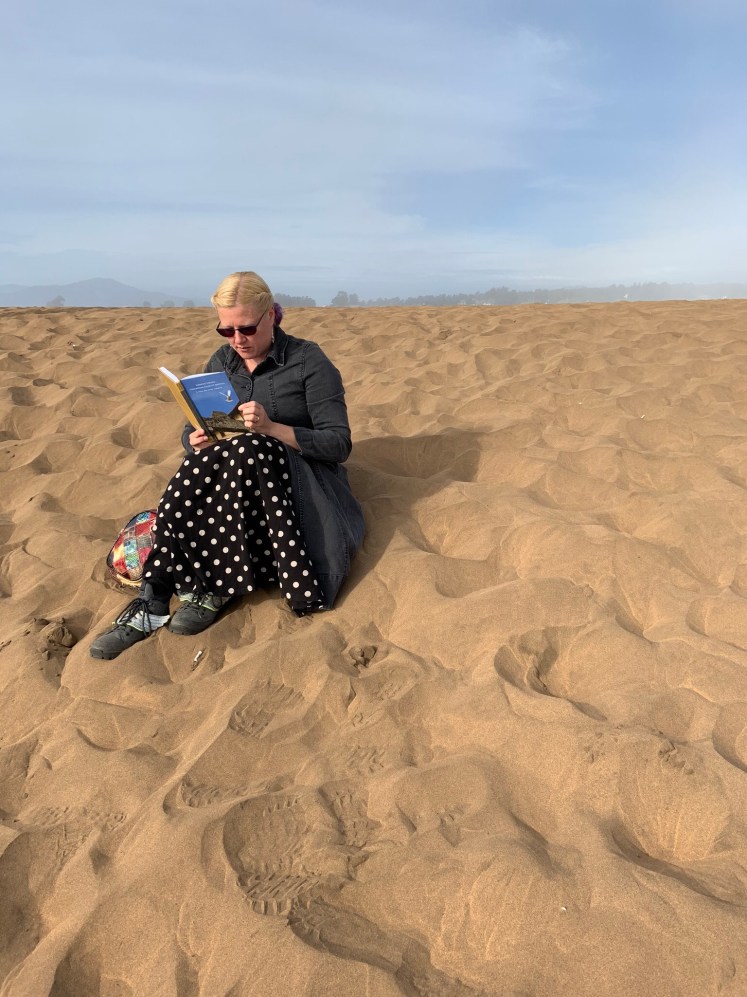 This is a photo of me sitting in the red sand and reading Narrating Violence, Constructing Collective Identities: 'To Witness These Wrongs Unspeakable' by Giti Chandra.