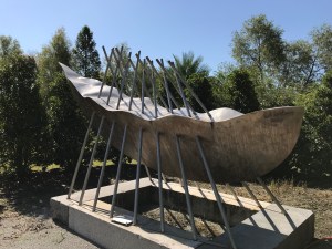 Picture of a long boat-shaped metal sculpture surrounded by trees.